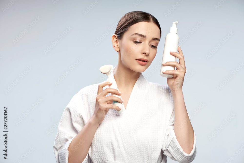 Attractive young lady holding bottle of cosmetic cream and pore cleansing brush Attractive young lady holding bottle of cosmetic cream and pore cleansing brush