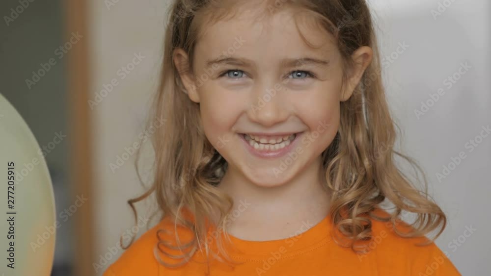 Little girl looks out from behind balloon and laughs happily. Close up portrait of a happy little child bursting in laugh. Child girl blonde with blue eyes and laughs indoors. Closeup. Slow Motion. Little girl looks out from behind balloon and laughs happily. Close up portrait of a happy little child bursting in laugh. Child girl blonde with blue eyes and laughs indoors. Closeup. Slow Motion.