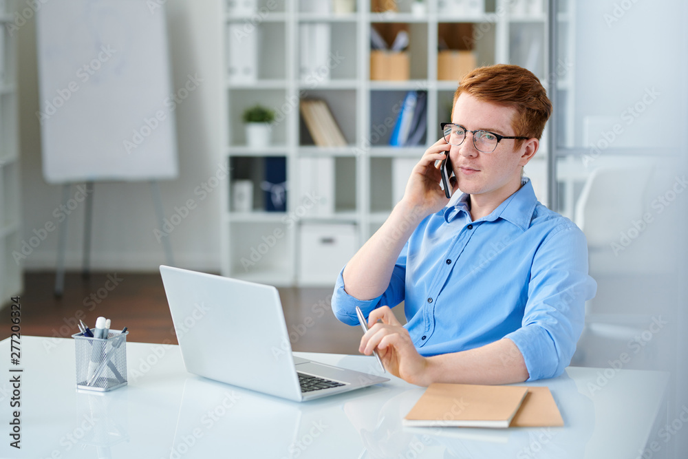 Young serious manager in blue shirt talking by smartphone by desk Young serious manager in blue shirt talking by smartphone by desk