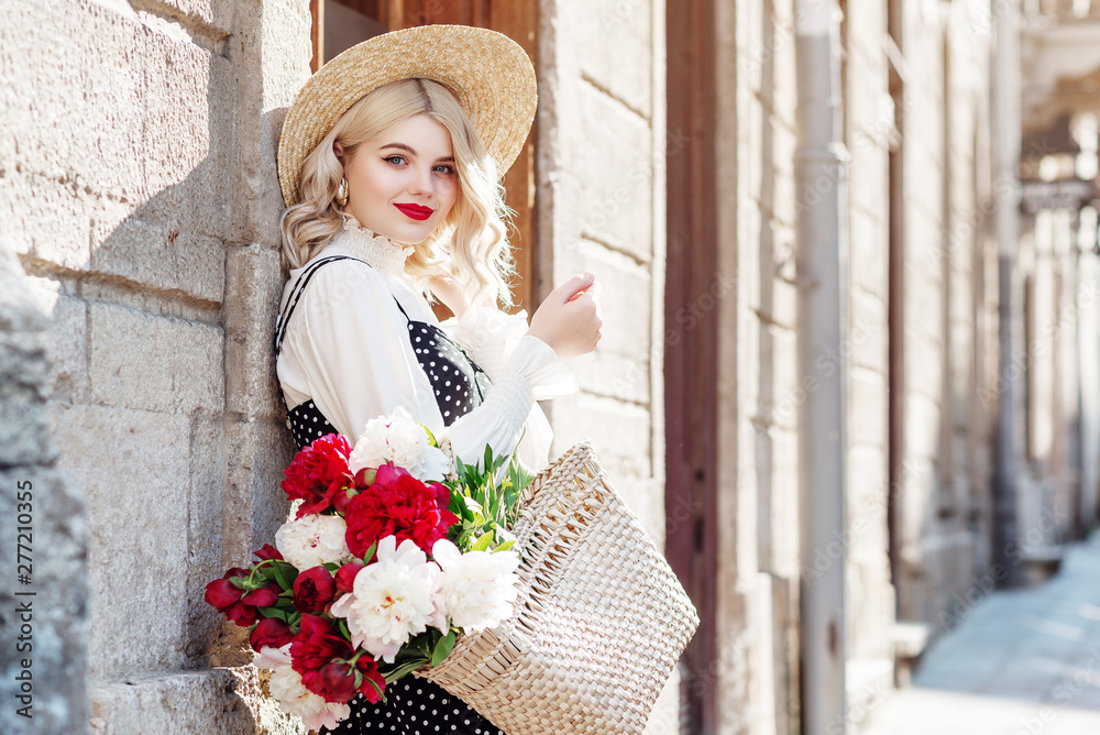 Outdoor portrait of young happy smiling lady wearing straw hat, white blouse, polka dot dress, holding wicker bag with peony flowers, posing in street of European city. Copy, empty space for text