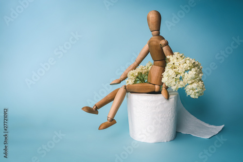 Wooden figure sit on a roll of toilet paper and flower of buldenege on a blue background. Toilet paper with a smell. Concept of the problem with digestion. photo