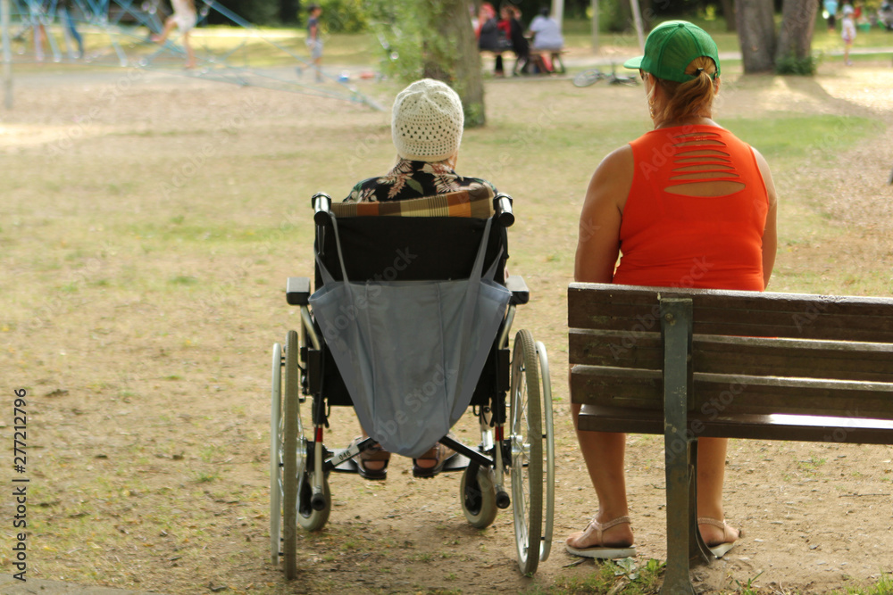 woman in an orange jacket and an old woman are disabled in a wheelchair, sit together in a park in summer woman in an orange jacket and an old woman are disabled in a wheelchair, sit together in a park in summer