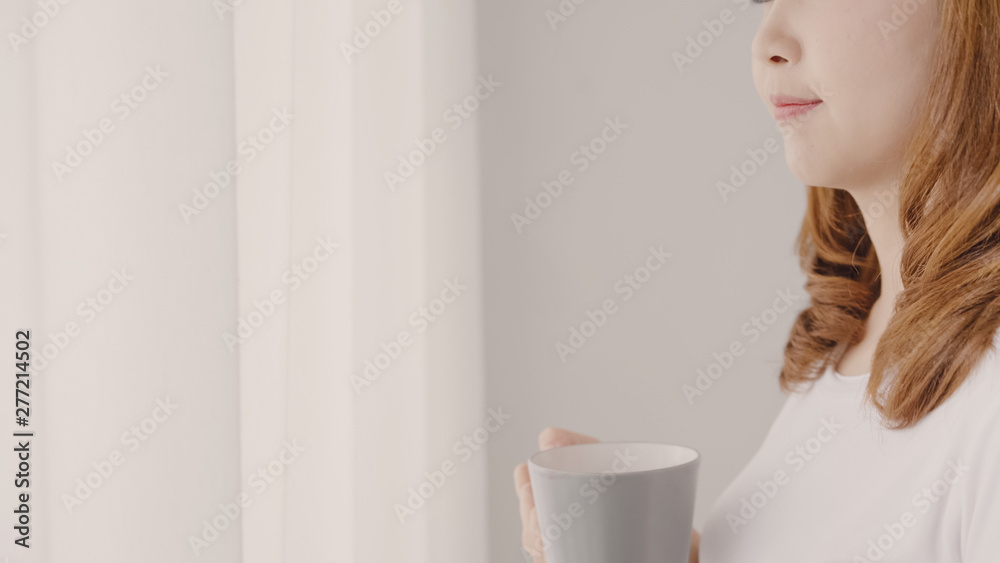 Happy beautiful Asian woman smiling and drinking a cup of coffee or tea near the window in the bedroom. Young asia female open the curtains and relax in the morning. Lifestyle woman at home concept. Happy beautiful Asian woman smiling and drinking a cup of coffee or tea near the window in the bedroom. Young asia female open the curtains and relax in the morning. Lifestyle woman at home concept.