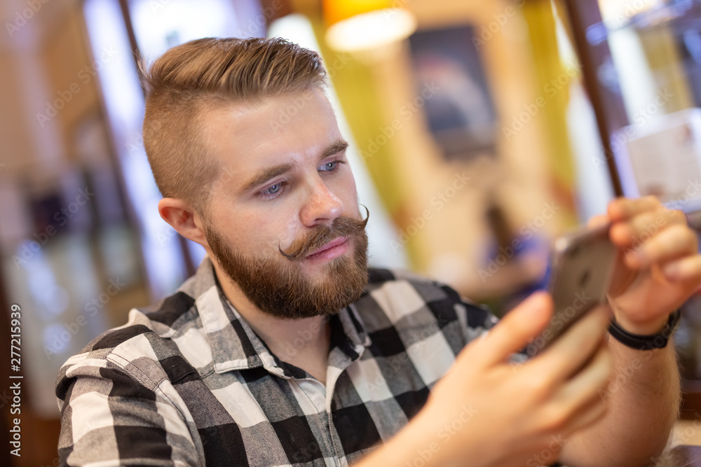Stylish young man with a mustache and beard is watching a social network using a smartphone while sitting in a cafe on a weekend. The concept of dependence on social networks. Stylish young man with a mustache and beard is watching a social network using a smartphone while sitting in a cafe on a weekend. The concept of dependence on social networks.