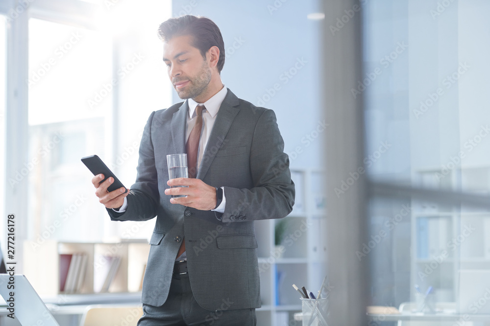 Young confident agent with glass of water scrolling in smartphone - obrazy, fototapety, plakaty 