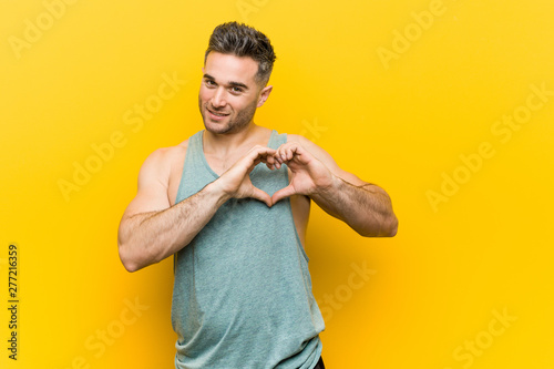 Young fitness man against a yellow background smiling and showing a heart shape with hands.