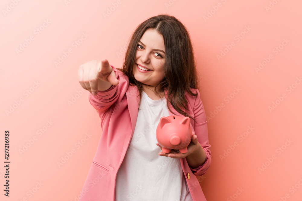 Young plus size curvy business woman holding a piggy bank cheerful smiles pointing to front. Young plus size curvy business woman holding a piggy bank cheerful smiles pointing to front.