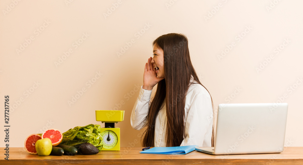 Young nutritionist chinese woman working with her laptop shouting and holding palm near opened mouth. Young nutritionist chinese woman working with her laptop shouting and holding palm near opened mouth.