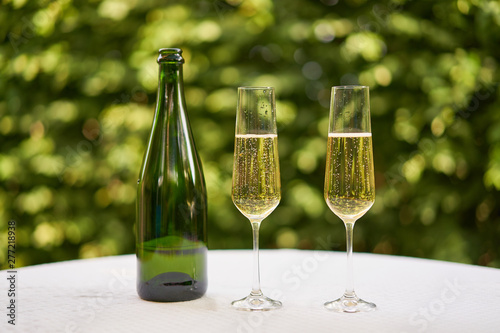 Two wine glasses flute type with sparkling wine or champagne and traditional design champagne bottle on the table with white tablecloth in the romantic garden restaurant ready to celebrate or cheers.