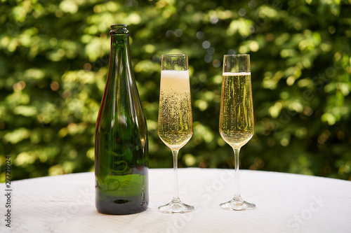 Close up Picture on two wine glasses flute type with sparkling wine or champagne and traditional design champagne bottle on the table with white tablecloth in the romantic garden restaurant, cheers.