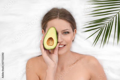Beautiful woman with avocado and tropical leaf on white fabric, above view. Organic face mask