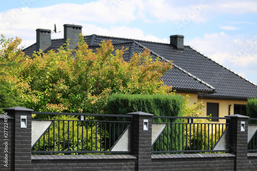 Murowane ogrodzenie z cegły i stali, dach budynku i zielone drzewa. 