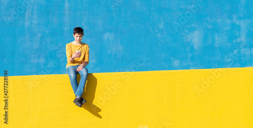 Front view of a young boy wearing casual clothes sitting on a yellow fence against a blue wall while using a smartphone