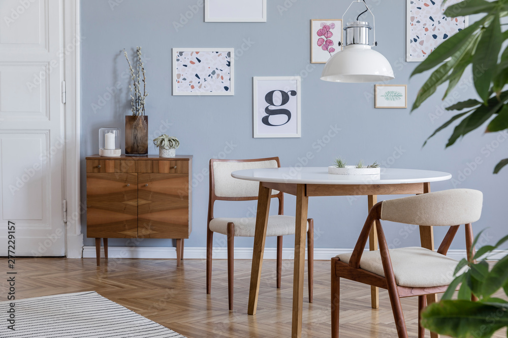 Fototapeta premium Stylish scandinavian home interior of sitting room with design wooden chairs, family table, plants, accessories and mock up posters gallery wall. Gray background walls. Modern home decor. Template.