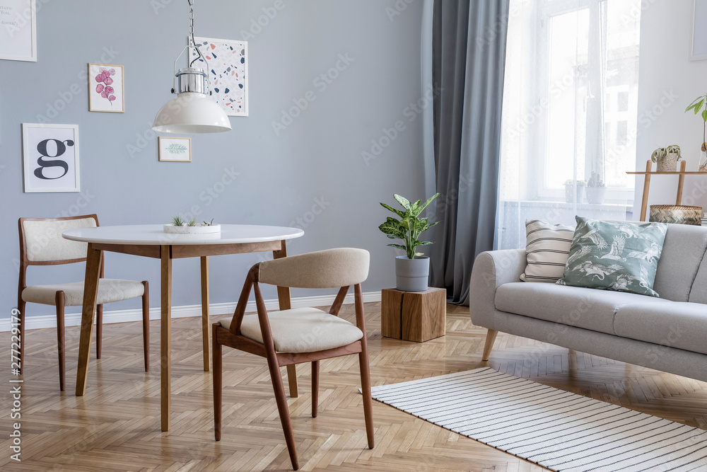 Design scandinavian home interior of open space with stylish chairs, family table wooden commode, gray sofa, accessories and mock up posters gallery wall. Gray background walls. Retro cozy home decor. Design scandinavian home interior of open space with stylish chairs, family table wooden commode, gray sofa, accessories and mock up posters gallery wall. Gray background walls. Retro cozy home decor.
