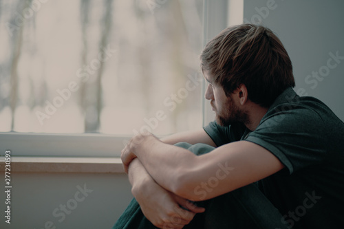 Fotografija Sad young man sitting on the floor looking through the window