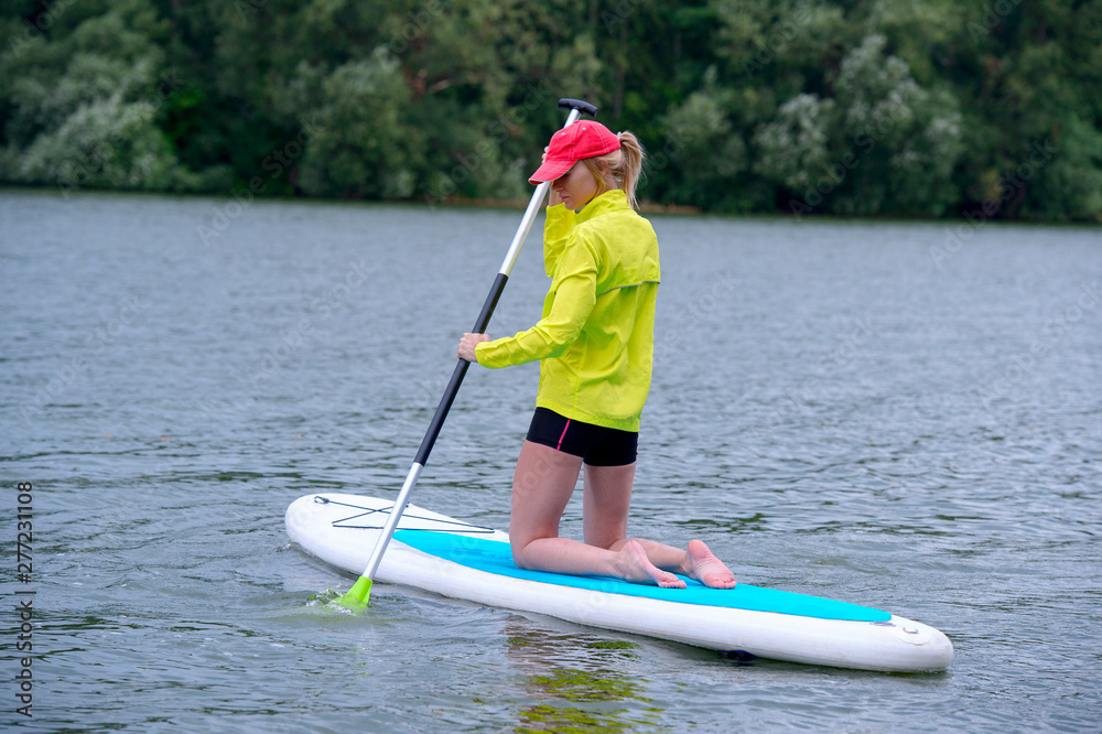 happy girl in a jacket is kneeling on a surfboard and holding a long paddle. Rear view photo