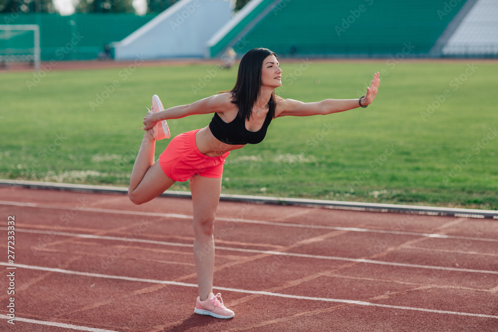Young brunette woman athlete in pink shorts and top on stadium sporty lifestyle standing stretching leg side view