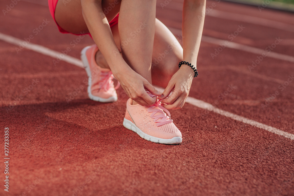 Young woman hands tie laces on her pink sport shoes on a stadium on a running path. White stripe near her. Close-up of tying shoes