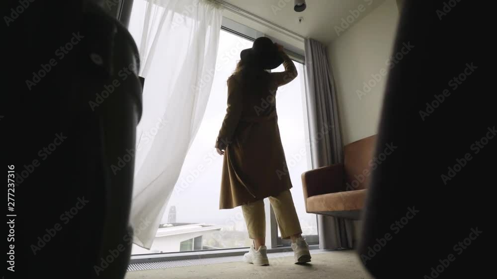 Back view silhouette woman just arrived in hotel apartment. low angle view girl coming to full length window, opening curtains throwing hat and enjoying view. Concept summer vacation 