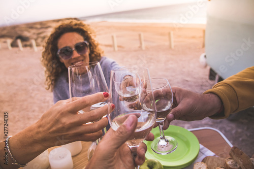 Happy friends cheering and drinking wine during camping vacation at the beach with vintage van. People having fun at weekend summer evening with camper. Youth lifestyle togetherness and party concept