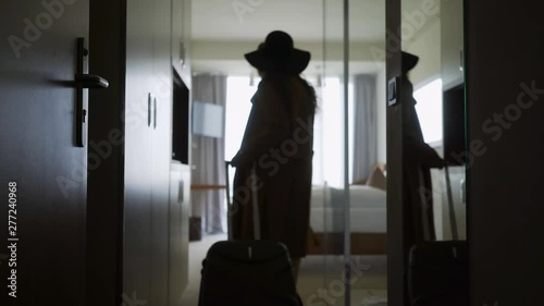 Back view woman opening door and entering in hotel apartment. Follow faceless female in hat with suitcase moving room, coming to full length window. Concept comfortable vacation