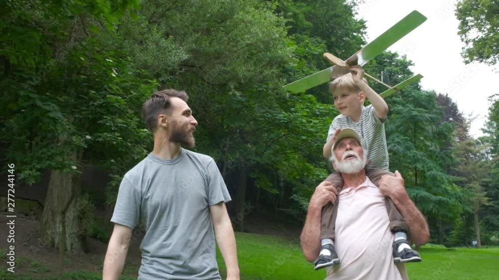 Little boy with airplane model and grandfather raising hands over green park on background enjoying life and nature. Portrait of happy grandfather giving grandson piggyback ride on his shoulders.