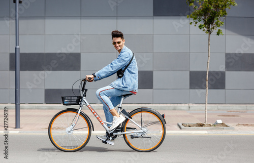 Stylish guy dressed in trendy jeans clothes rides bicycle in the street against the background of a gray building