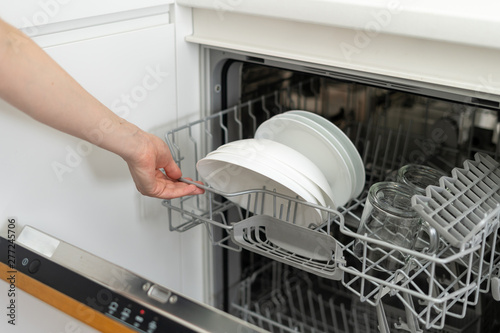 Woman opening modern dishwasher machine with plates and cups, standing on kitchen