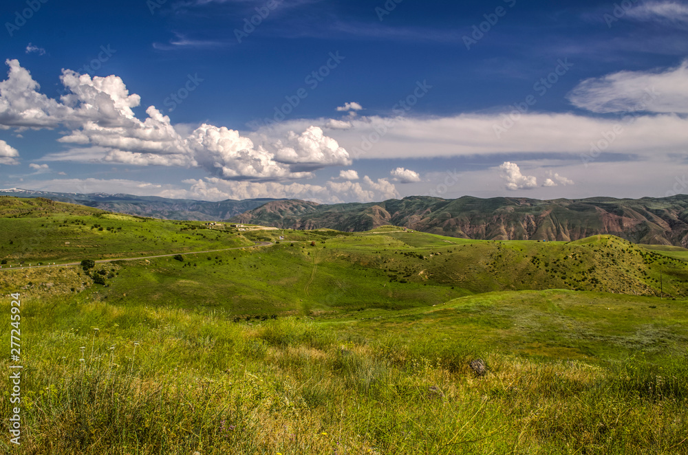 View of the highway with a turn leading to the Garni temple among the mountains of Geghama ridge in Armenia against the blue sky covered with huge clouds - obrazy, fototapety, plakaty 