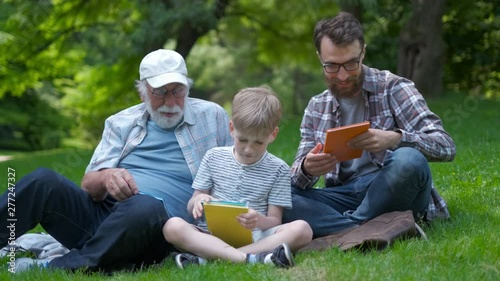 happy family of three generation - father, grandfather and blond son sitting on grass at park with books learn to read while getting ready for school.They are laugh,fun,spending good time together photo