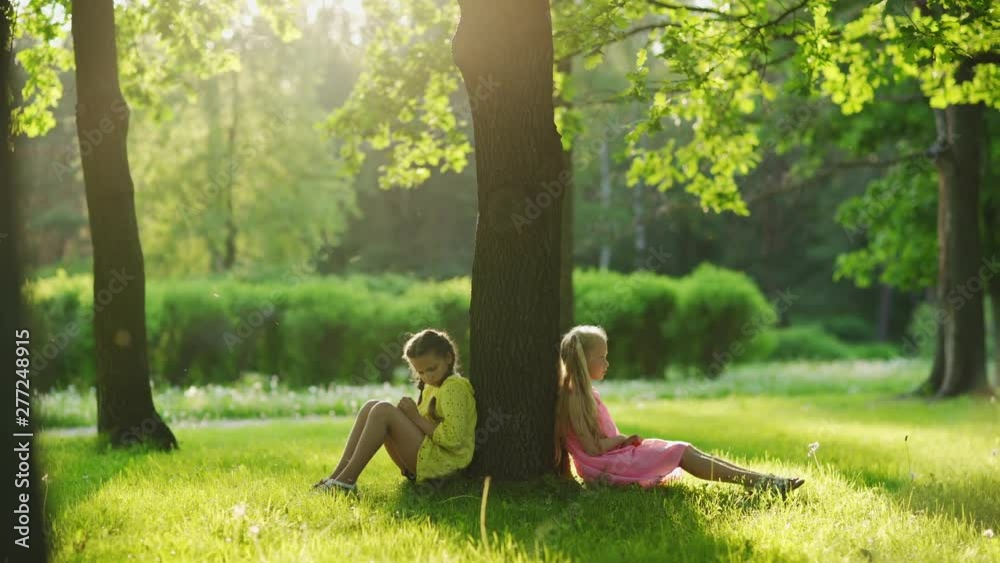 Two sulking little girls sitting on grass in park, leaning on tree trunk back to back and worrying after best friends quarrel on sunny summer day, tracking right shot