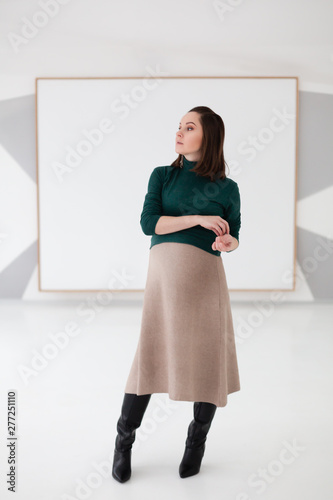 Bild auf Leinwand Young and beautiful pregnant fashionable woman in beige skirt and green sweater indor on white background touching her hand