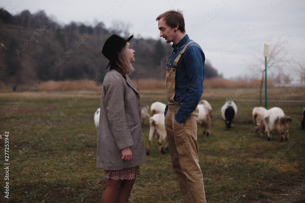 Love story. Woman in hat and her boyfriend standing next to each other at the field full of goats somewhere at countryside Love story. Woman in hat and her boyfriend standing next to each other at the field full of goats somewhere at countryside