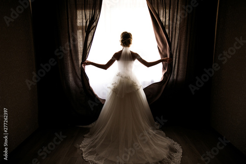 the bride in a wedding dress with a train opens the curtains of the window, stands back, you can see her silhouette, against the light