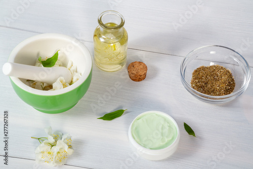Jar of cream made from natural plant ingredients, oils and herbs, jasmine flowers, mortar and pestle on a white wooden background - preparation of organic cosmetics concept