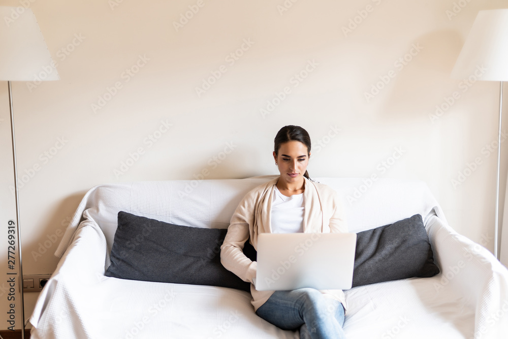 Young woman doing research work for her business. Smiling woman sitting on sofa relaxing while browsing online shopping website. Happy girl browsing through the internet during free time at home. Young woman doing research work for her business. Smiling woman sitting on sofa relaxing while browsing online shopping website. Happy girl browsing through the internet during free time at home.