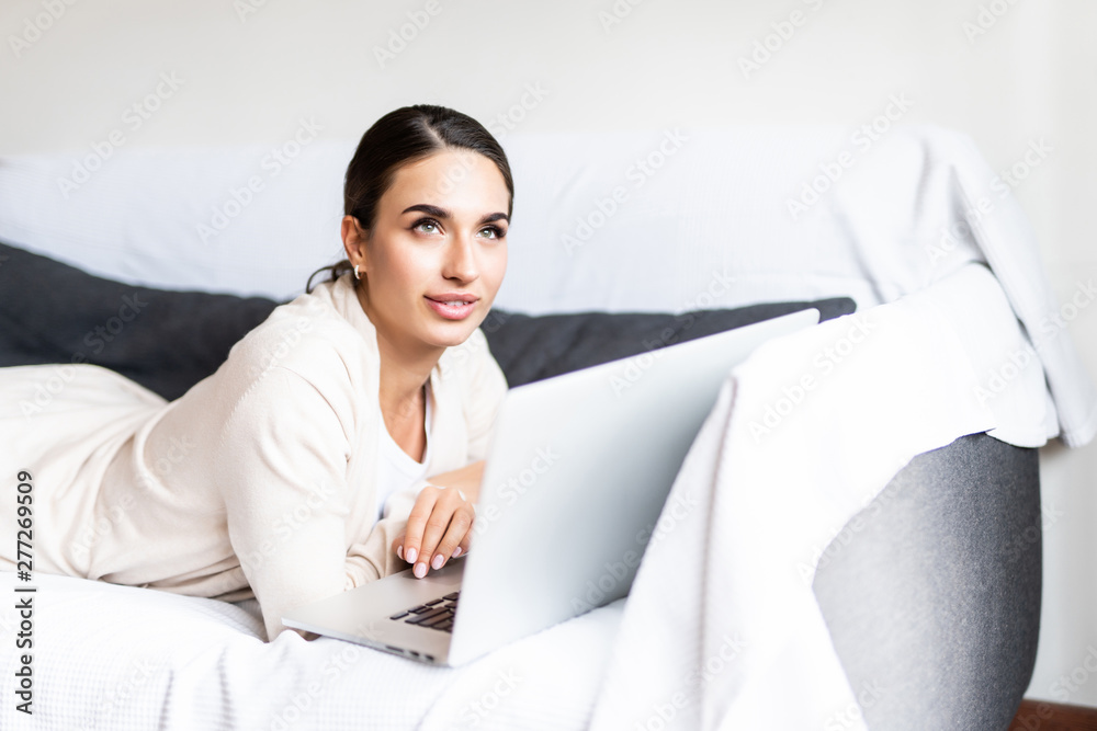 Smiling young woman lying on sofa with laptop in front of her in the living room Smiling young woman lying on sofa with laptop in front of her in the living room