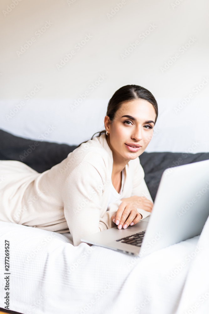 Smiling young woman lying on sofa with laptop in front of her in the living room Smiling young woman lying on sofa with laptop in front of her in the living room