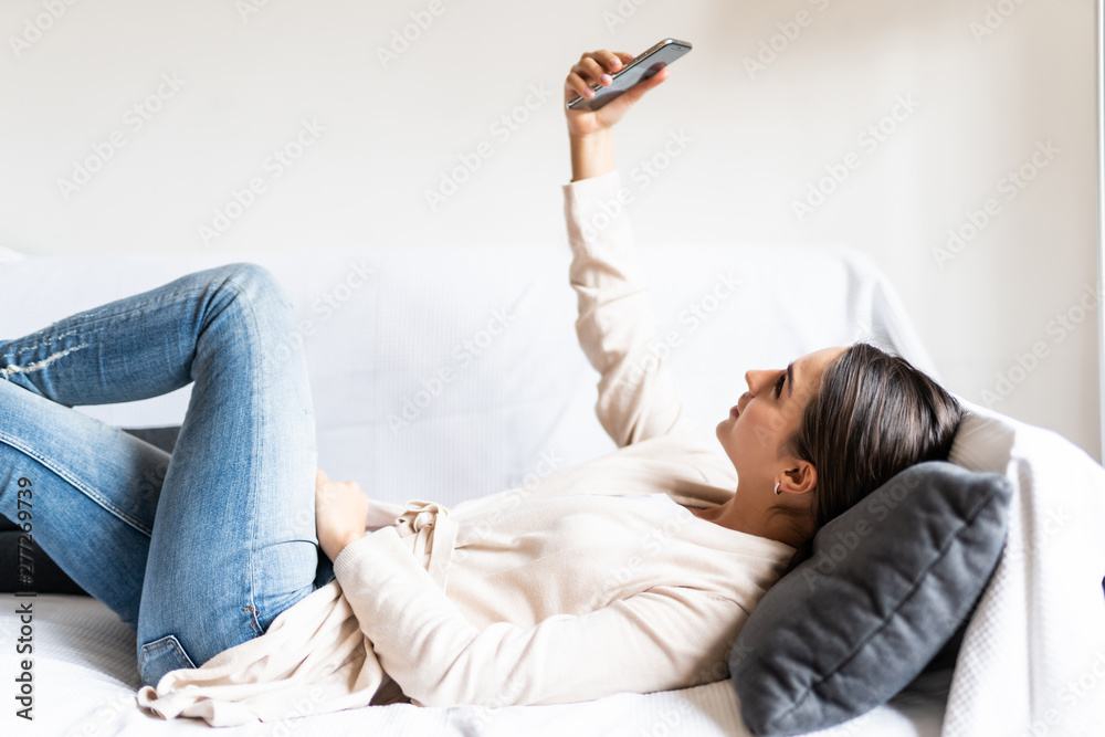 Photo of brunette woman taking selfie on cell phone while sitting on couch in bright apartment Photo of brunette woman taking selfie on cell phone while sitting on couch in bright apartment