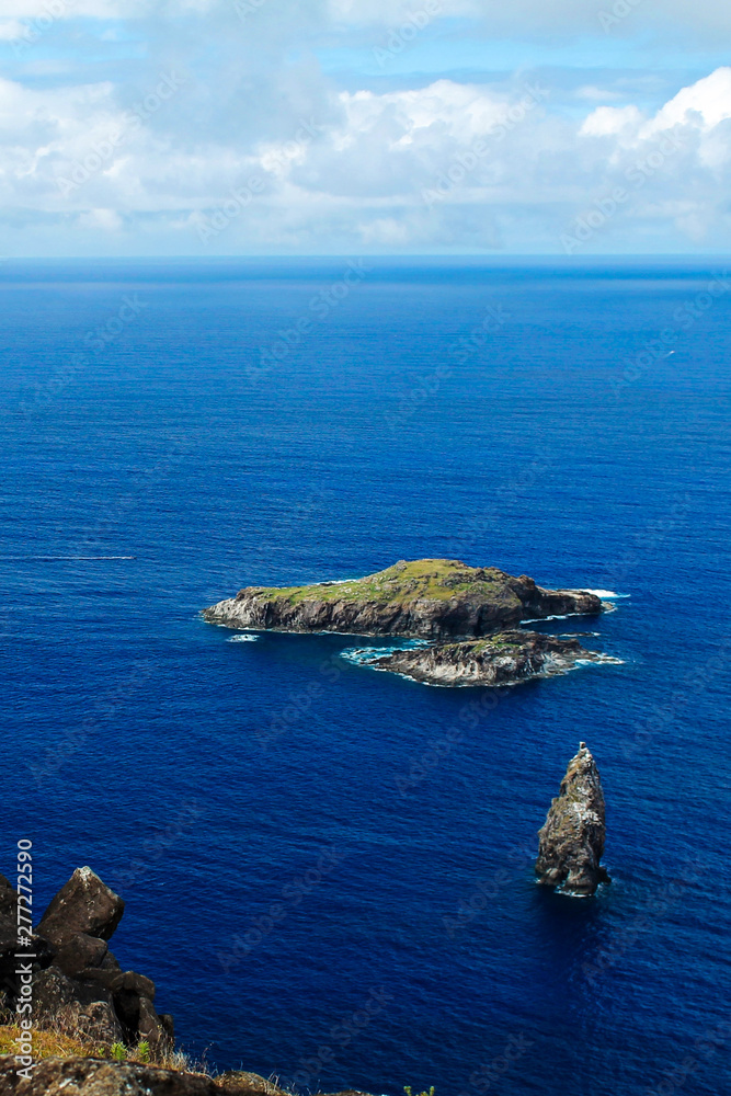 View of the motus (islets) off the coast of Easter Island, Chile, captured from a cliff top, showcasing the deep blue tones of the Pacific Ocean on a sunny day