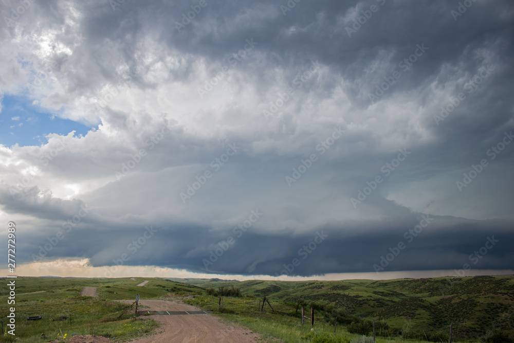 A supercell storm cloud hangs low in the sky over the prairie and a dirt road extends to the horizon.