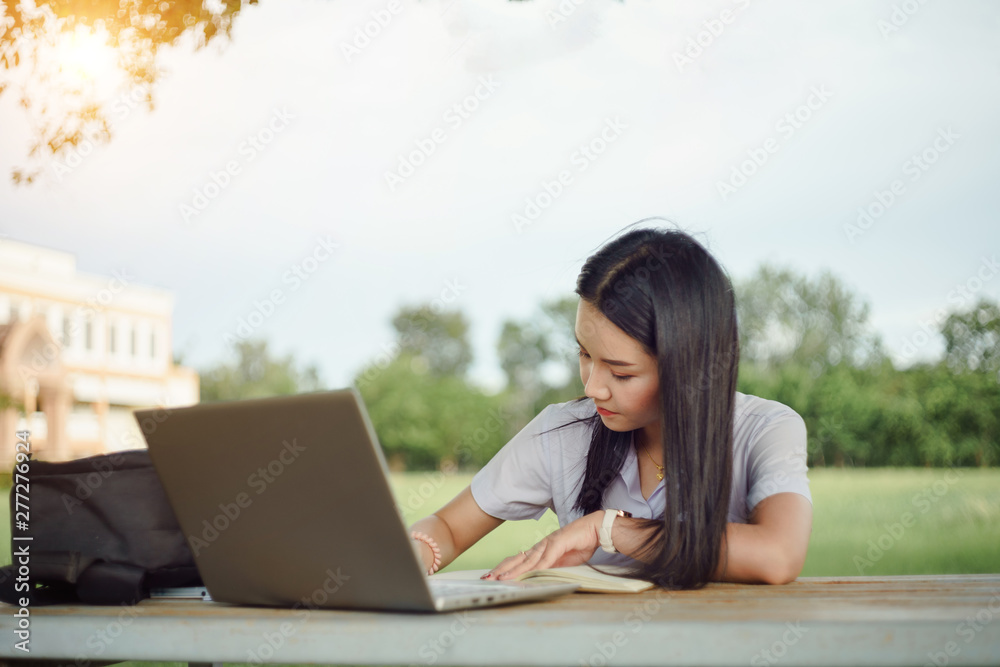 A young female university student writes a note and assignment with a laptop beside outside the campus with blure green tree background. Students self learning outside the campus. A young female university student writes a note and assignment with a laptop beside outside the campus with blure green tree background. Students self learning outside the campus.