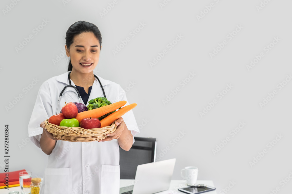 A young woman doctor nutritionist smiling siting working on her laptop in her office holding a basket of vegetables. A young woman doctor nutritionist smiling siting working on her laptop in her office holding a basket of vegetables.
