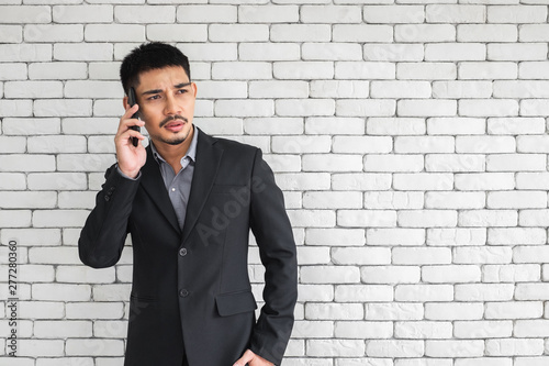 An Asian man in business suit on mobile phone and looking concern, standing in front of white brick wall.