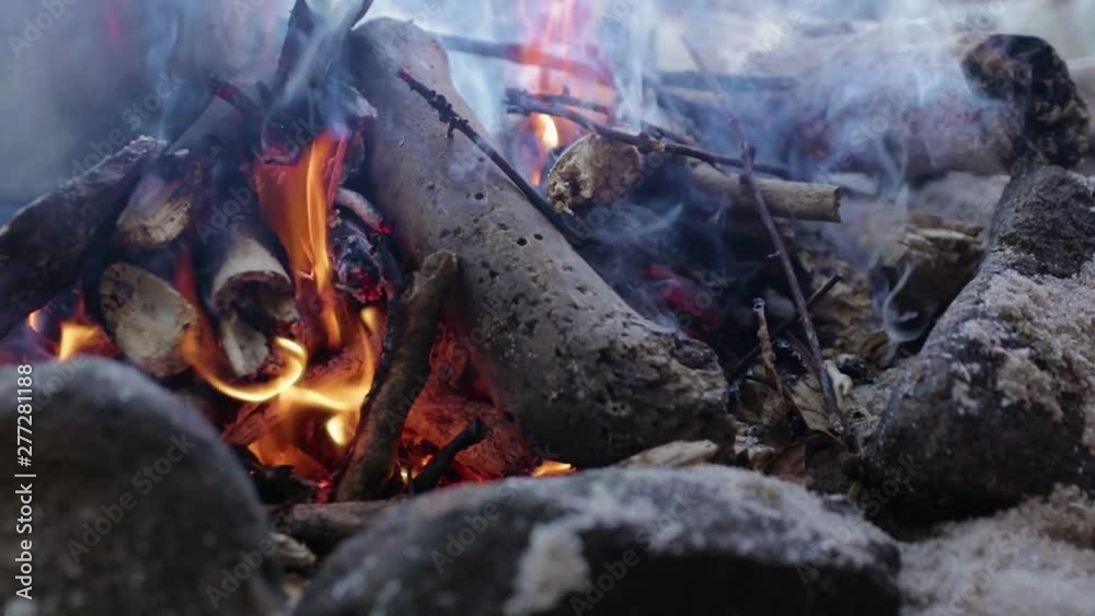 Close up of fire in a camp during a sunny day ate the beach with strong and beautiful colors, some sand in the background, and in slow motion Close up of fire in a camp during a sunny day ate the beach with strong and beautiful colors, some sand in the background, and in slow motion