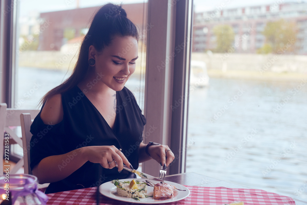 Brunette girl with a tail sitting in a restaurant on ship on the background of the river. Girl tries salmon with rice. Girl eats appliances. Woman with the tunnels in the ears and piercing in the nose