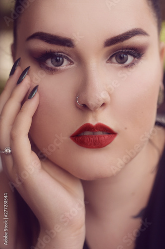 Portrait of a girl with professional make-up .smoky eyes  red lips and smooth eyebrows. Limbed eyebrows. Girl with piercing in the nose. Piercing  rings. Brunette with long nails  manicure.