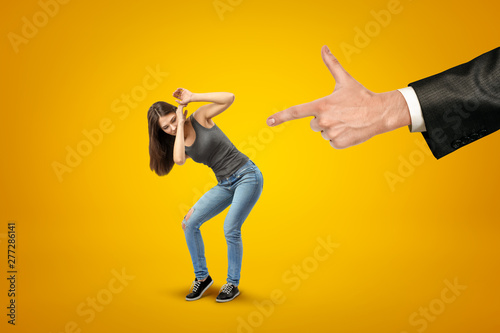 Man's hand in suit pointing fingergun at miniatured pretty girl in casual clothes, who is protecting her face with hands