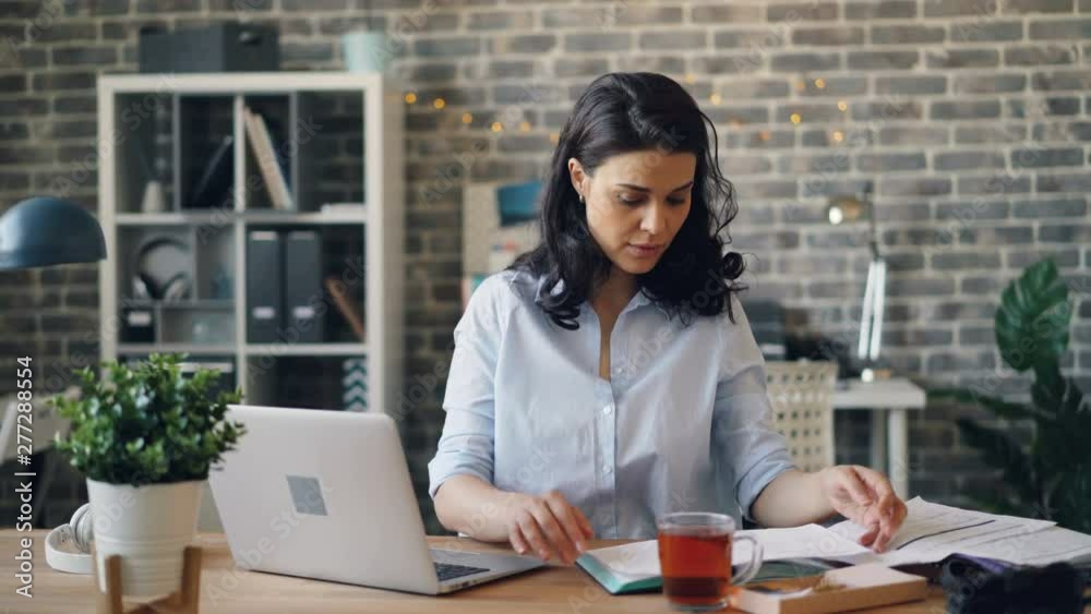 Worried manager pretty lady is using laptop and reading paper document working in office alone with sad face. Problems at work and modern business concept.